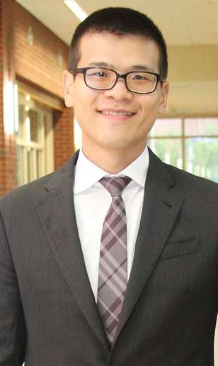 Chung-Hao Lee has short hair and glasses and is smiling. He is wearing a dark gray suit jacket, white collared dress shirt, and a plaid tie, conveying a professional demeanor. The background shows a hallway with windows and red brick walls.